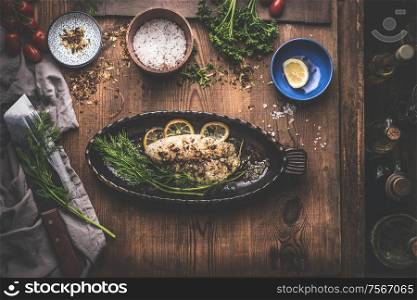 Fish fillet with lemon and herbs in backing pan on rustic kitchen table background with ingredients. Top view
