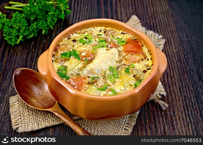 Fish soup with tomatoes, potatoes, pepper and cream in a clay bowl on burlap, parsley on a background of a dark wooden board