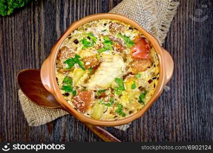 Fish soup with tomatoes, potatoes, peppers and cream in a clay bowl on a sackcloth napkin, parsley on a wooden board background from above