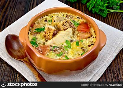 Fish soup with tomatoes, potatoes, peppers and cream in a clay bowl on a napkin, parsley on a dark wooden board background