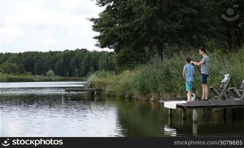 Fisherman checking the bait and tackle of spinning rod and reel while fishing together with teenage son at freshwater pond on summer day. Positive hipster dad and boy with fishing rods angling on the lake against colorful rural landscape background.