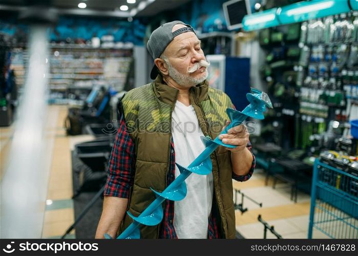 Fisherman holds a drill for winter fishing in shop. Male angler buying equipment and tools for fish catching and hunting, assortment on showcase in store. Fisherman holds a drill for winter fishing in shop