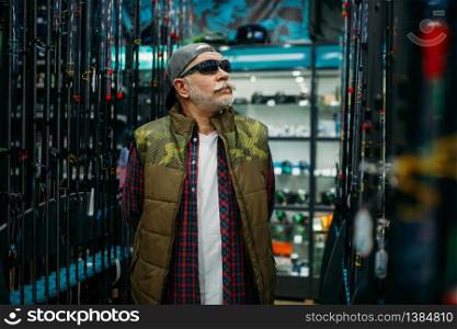 Fisherman in sunglasses choosing rod in fishing shop. Equipment and tools for fish catching and hunting, accessory choice on showcase in store. Fisherman choosing rod in fishing shop