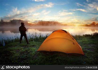 Fisherman near an orange tent on the bank of a misty river at sunset. Fisherman near an orange tent on bank of misty river at sunset