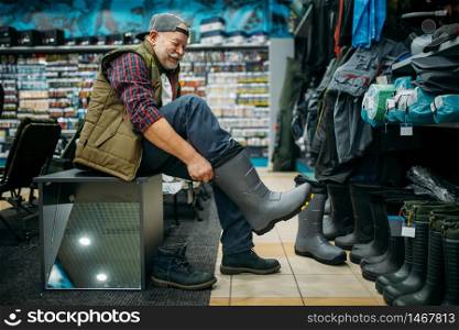 Fisherman tries on rubber boots in fishing shop. Equipment and tools for fish catching and hunting, accessory choice on showcase in store. Fisherman tries on rubber boots in fishing shop