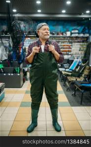 Fisherman tries on rubber jumpsuit in fishing shop, hooks and baubles on background. Equipment and tools for fish catching and hunting, accessory choice on showcase in store. Fisherman tries on rubber jumpsuit in fishing shop
