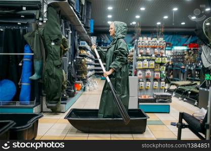 Fisherwoman in a raincoat is sailing on a boat in fishing shop, side view, hooks and baubles on background. Fisherwoman shoosing equipment and tools for fish catching and hunting, accessory in store. Fisherwoman is sailing on a boat in fishing shop