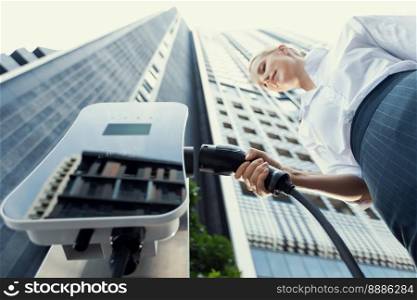 Fisheye view focus hand holding, inserting, plugging ev charger from public charging station for electric vehicle with blurred progressive businesswoman and residential city building in background.. Fisheye view progressive businesswoman plugs charger from charging station to EV