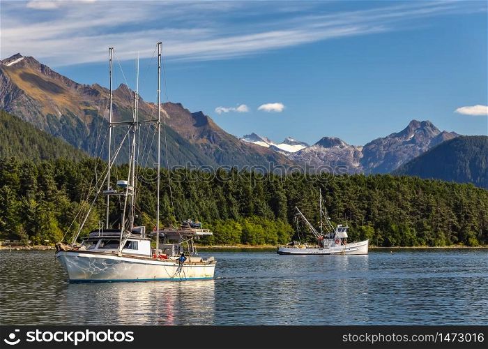 Fishing boat sailing in harbor and another fishing boat drifting behind it. Mountains, forest and blue sky in the background. Sitka, Alaska, USA