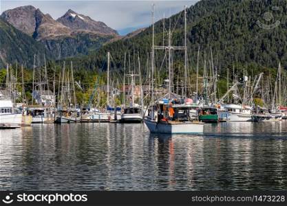 Fishing boat sailing in marina and is about to dock. Marina full of other fishing boats. Gorgeous mountains and forest in the background. Sitka, Alaska, USA