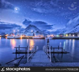 Fishing boats near pier on the sea against snowy mountains and starry sky with moon at night in Lofoten islands, Norway. Winter landscape with bridge, ship, buildings, illumination, rocks and clouds