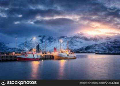 Fishing boats on the sea, snowy mountains, colorful sky with clouds at sunset in Lofoten islands, Norway. Winter landscape with boats, harbor, lights, houses with illumination, rocks in snow. Nature