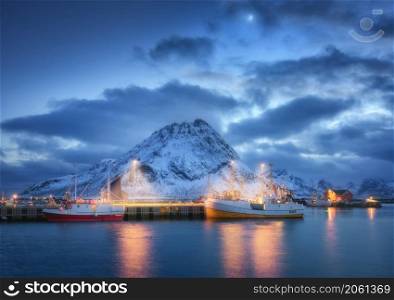 Fishing boats on the sea, snowy mountains, sky with clouds, moon at night in Lofoten islands, Norway. Winter landscape with boats, harbor, city lights, houses with illumination, rocks in snow. Nature