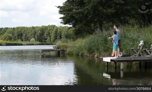 Fishing by the lake is our common passion. Happy father and excited son fishing with spinning rod on wooden pier by the pond in summer over amazing landscape background. Carefree family enjoying time together fishing on the lake.
