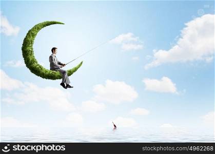 Fishing concept. Businessman sitting on green moon and fishing with rod