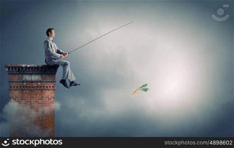Fishing concept. Businessman sitting on top of building and fishing with rod