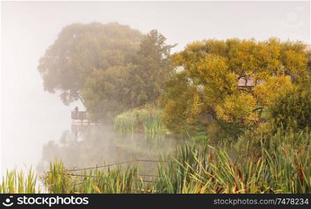 Fishing on foggy lake. Fisherman with rods on a lake, tranquil water surface, reflection and deep fog on a cold autumn morning, Belarus