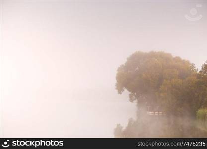 Fishing on foggy lake. Fisherman with rods on a lake, tranquil water surface, reflection and deep fog on a cold autumn morning, Belarus