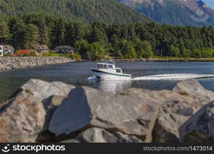 Fishing speedboat sailing fast in harbor. Long water trace behind it. Rocks in soft focus in the foreground. Houses, mountains, and forest in the background. Sitka, Alaska, USA
