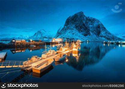 Fishing village with boats on the sea at night and snowy mountains in Lofoten islands, Norway. Winter landscape with houses, pier, illumination, rocks, moody sky reflected in water. Norwegian rorbu