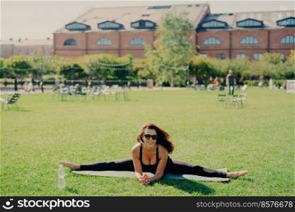 Fit active European woman does pilates on fitness mat wears sunglasses and sportswear shows great progress poses on green lawn has happy expression trains outside. Fitness and stretching concept