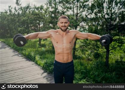 Fit adult man lifts heavy weight during workout session outdoor, poses with naked torso, has well shaped muscular body, demonstrates his power, lifts barbells. Motivated bodybuilder at street