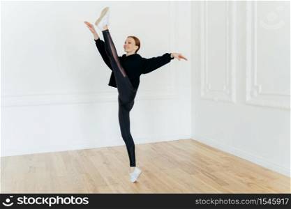 Fit athtletic woman demonstrates resiliance, balances on one leg, being skilled sport gymnast, dressed in sportswear and sneakers, has pilates exercises, stands in empty hall, has beautiful figure