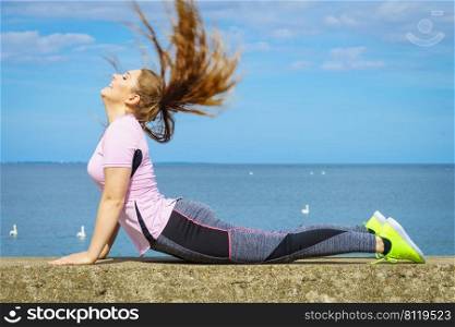 Fit attractive young woman wearing fashionable outfit working out being active outside. Practice yoga next to sea, having windblown hair.. Woman doing yoga next to sea