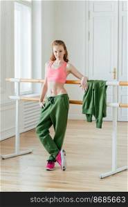 Fit ballet dancer in class near barre, rests on bar, has combed ginger hair, wears top and sport trousers, stands crossed legs in fitness centre, warms up before practice starts. Choreography concept