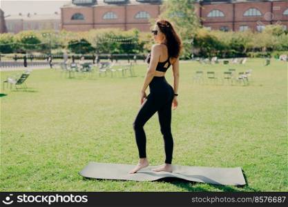 Fit brunette woman dressed in sportswear stands bare feet on karemat has fintess training on green lawn outdoor during summer time wears sunglasses looks somewhere being in good physical shape