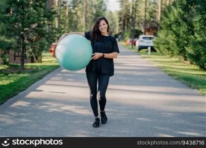Fit brunette woman exercises with fitness ball, dressed in active wear, poses, walks, and enjoys fresh air in the park. Embracing people, gymnastics, and aerobics concept.
