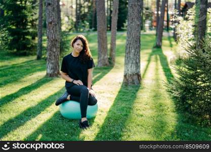 Fit brunette young woman takes rest after doing exercises at fitness ball leads active sporty lifestyle poses in urban environment on green grass against trees with sunshine does gymnastics.