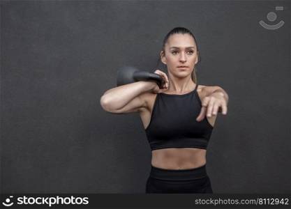 Fit female athlete in sportswear stretching out arm and holding heavy kettlebell on shoulder during functional training against black wall in gym. Determined sportswoman exercising with kettlebell