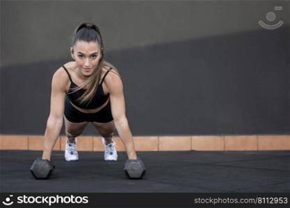 Fit female athlete in sportswear with ponytail doing plank exercise on dumbbells and looking at camera against black wall during fitness workout in gym. Sportswoman doing plank on dumbbells