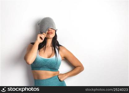 Fit female model dressed in sports bra and leggings covering half of face with beanie while standing with hand on waist on white background. Serious woman in sportswear covering face with hat