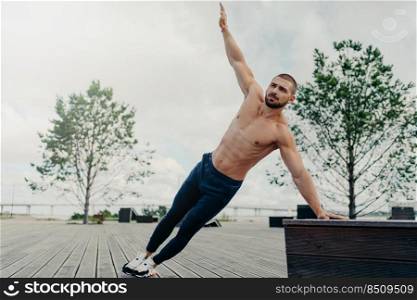 Fit healthy muscular man stands in side plank and raises arm, poses with naked torso, dressed in sport trousers and sneakers, exercises abs, raises abdominal crunch, trains outdoor near beach