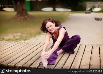 Fit healthy young woman exercising on a jetty. Fit healthy young woman exercising on a jetty over a lake or river smiling at the camera as she does stretching exercises holding her bare feet