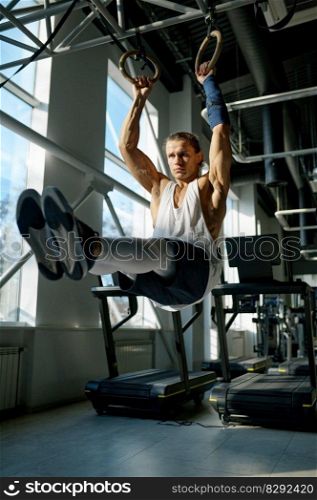 Fit male athlete training on gymnastics rings in light sport hall at gym. Muscular man with strong arms doing exercise lifting legs while hanging on sports equipment. Fit male athlete training on gymnastics rings in light sport hall at gym