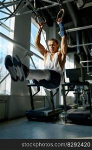 Fit male athlete training on gymnastics rings in light sport hall at gym. Muscular man with strong arms doing exercise lifting legs while hanging on sports equipment. Fit male athlete training on gymnastics rings in light sport hall at gym