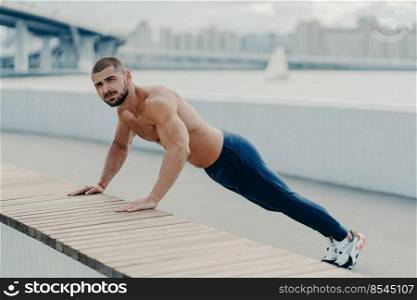 Fit muscular man does plank push up exercise leads healthy lifestyle wears sport trousers and sneakers poses outdoor near bridge. Motivated sportsman pushing hard. Sport, motivation and determination