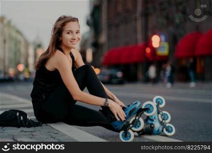 Fit positive woman puts on rollerblades sits on asphalt against blurred street background has positive expression goes in for sport has outdoor fitness activities during summer time. Hobby concept