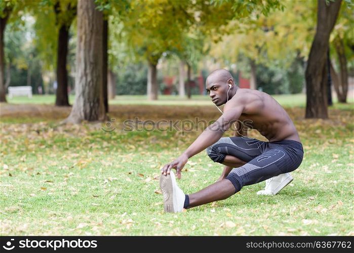 Fit shirtless young black man doing stretching before running in urban park. Young male exercising with naked torso listening to music with headphones.