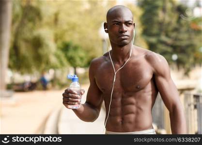 Fit shirtless young black man drinking water after running in urban background. Young male exercising with naked torso listening to music with headphones.