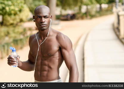 Fit shirtless young black man drinking water after running in urban background. Young male exercising with naked torso listening to music with headphones.