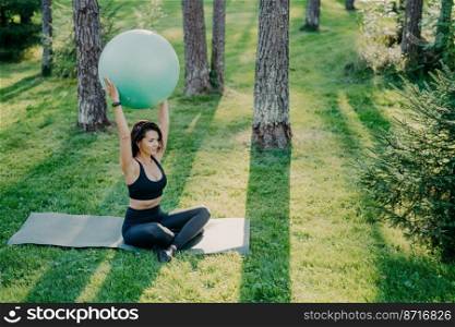 Fit sporty woman exercises with fitness ball sits on karemat in lotus pose dressed in cropped top and leggings, poses in forest during sunny day, breathes fresh air. Active lady practices yoga outside