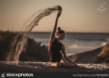 Fit woman in bodysuit sitting on beach of shoreline and pouring heap of sand making flying trail . Slim woman sitting on beach with sand trail