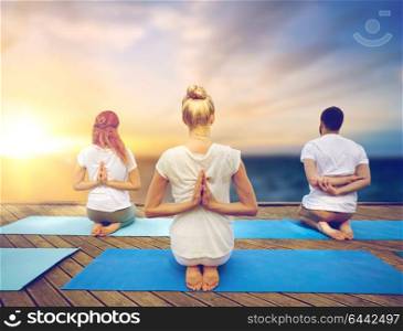 fitness and healthy lifestyle concept - group of people doing yoga reverse prayer pose on wooden pier over sea background. group of people doing yoga outdoors
