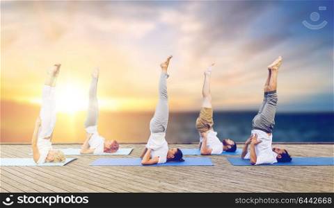 fitness and healthy lifestyle concept - group of people doing yoga supported shoulderstand pose on mat outdoors on wooden pier over sea background. people doing yoga shoulderstand on mat outdoors