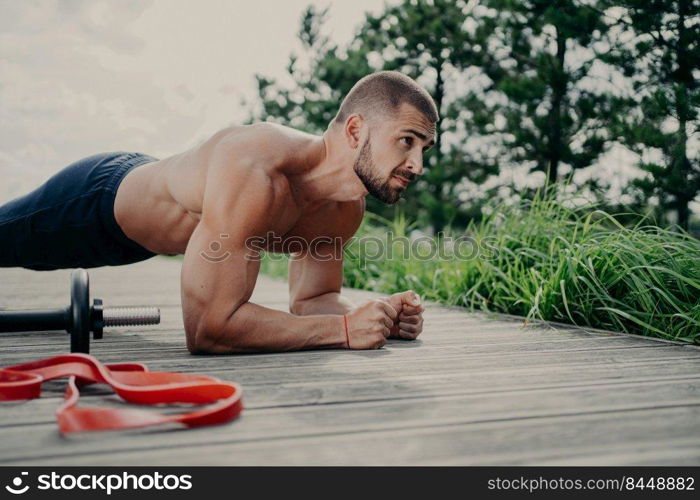 Fitness bearded Caucasian man with naked torso stands in plank pose, exercises outdoor, poses near sport equipment, has strong arms, demonstrates endurance and motivation. Abdominal workout.