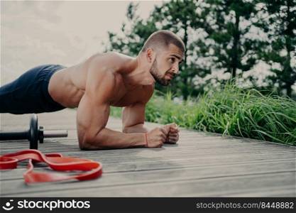 Fitness bearded Caucasian man with naked torso stands in plank pose, exercises outdoor, poses near sport equipment, has strong arms, demonstrates endurance and motivation. Abdominal workout.